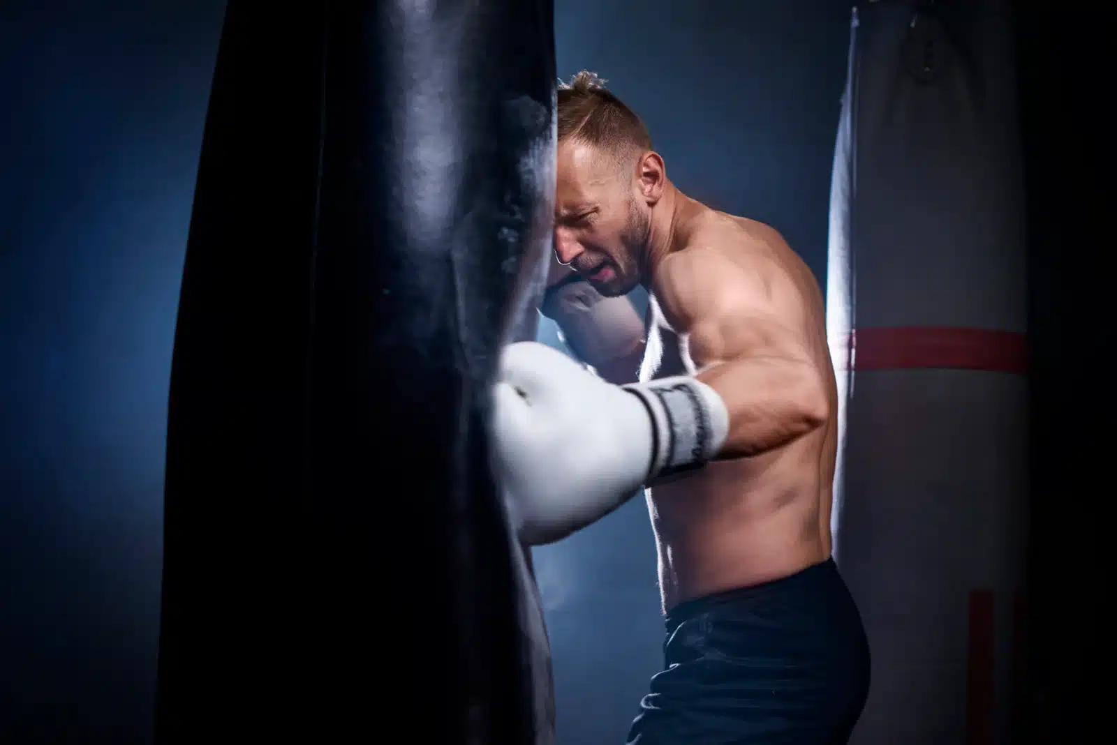 Boxeur Puissant Entraînement Sac de Frappe Intense Boxeur torse nu, le visage crispé par l'effort, frappant violemment un sac de frappe noir avec un gant blanc.