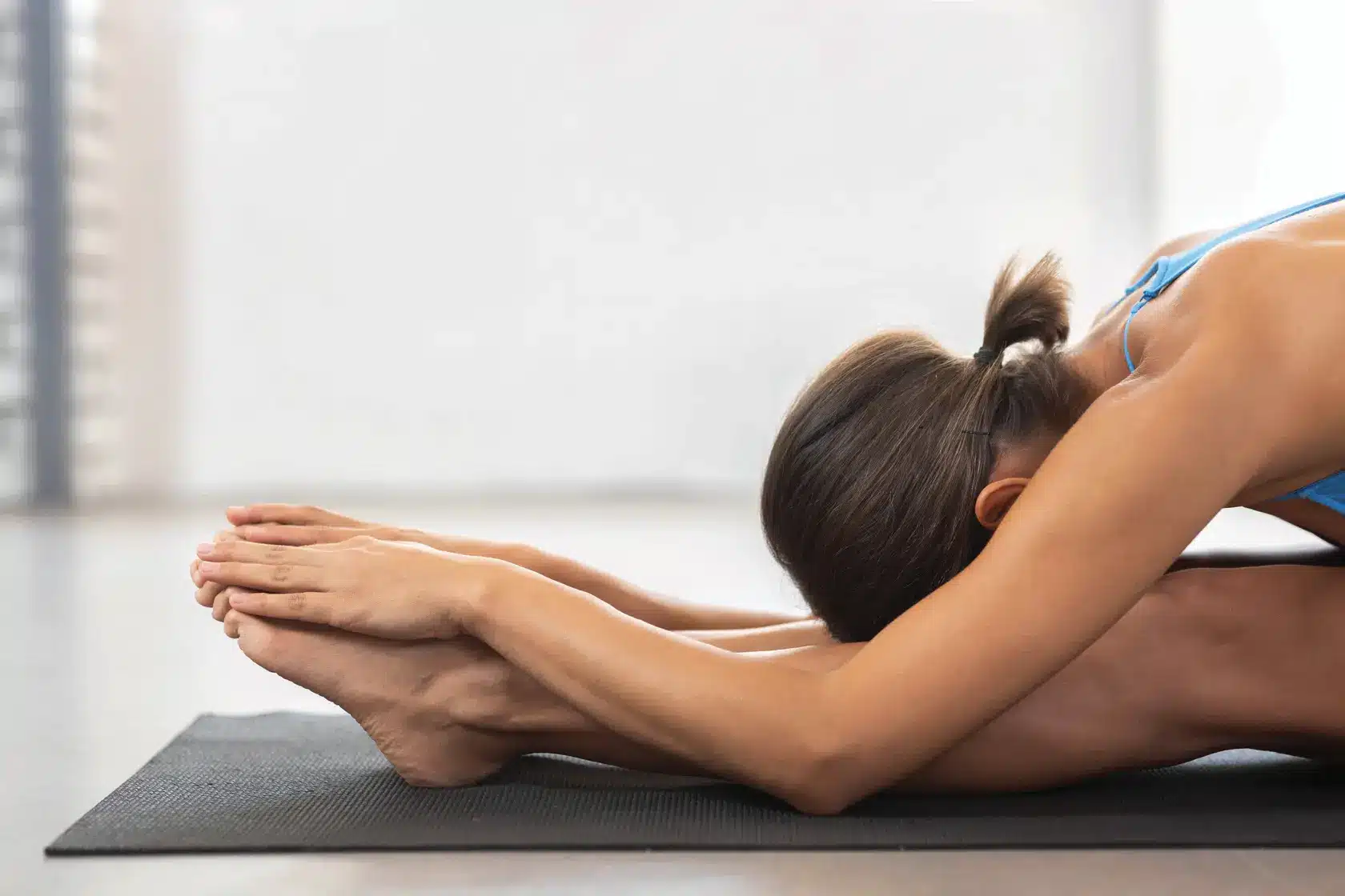 Jeune femme brune en haut bleu effectue un étirement assis, se penchant vers l'avant pour saisir ses pieds sur un tapis de yoga noir.