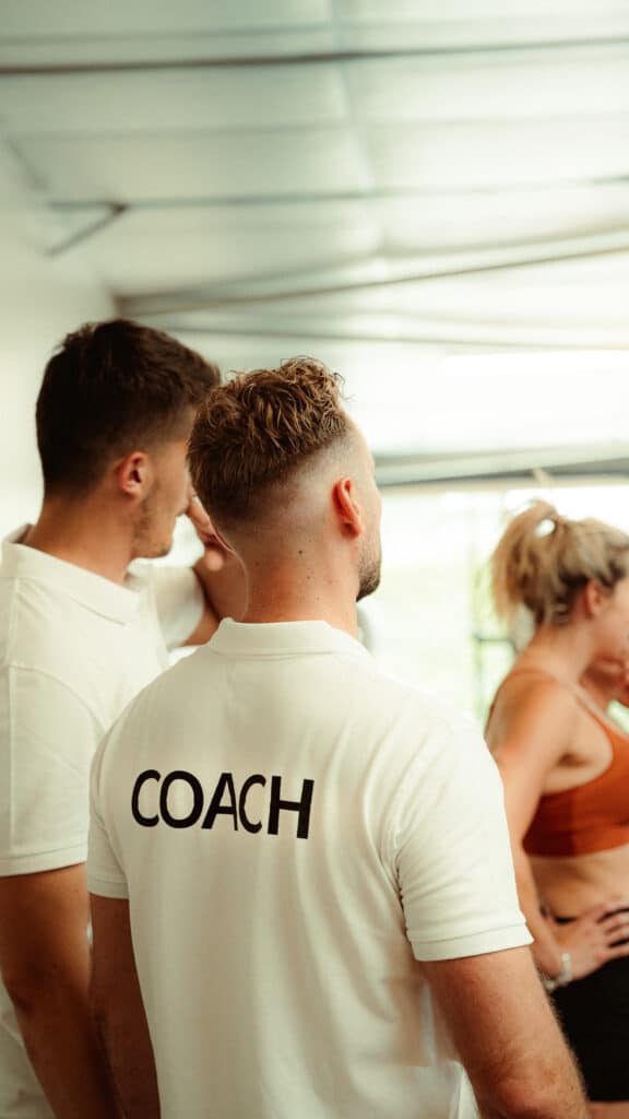 Coach sportif de dos, en polo blanc frappé du mot "COACH", dirigeant un groupe de sportifs dans une salle lumineuse.