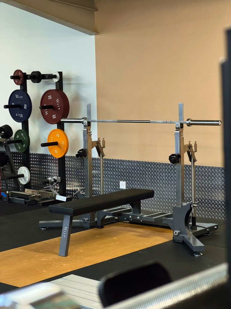 Banc de Musculation Witt et Équipement Haltérophilie Banc de musculation Witt avec barre olympique et rack de disques de poids colorés dans une salle de sport.