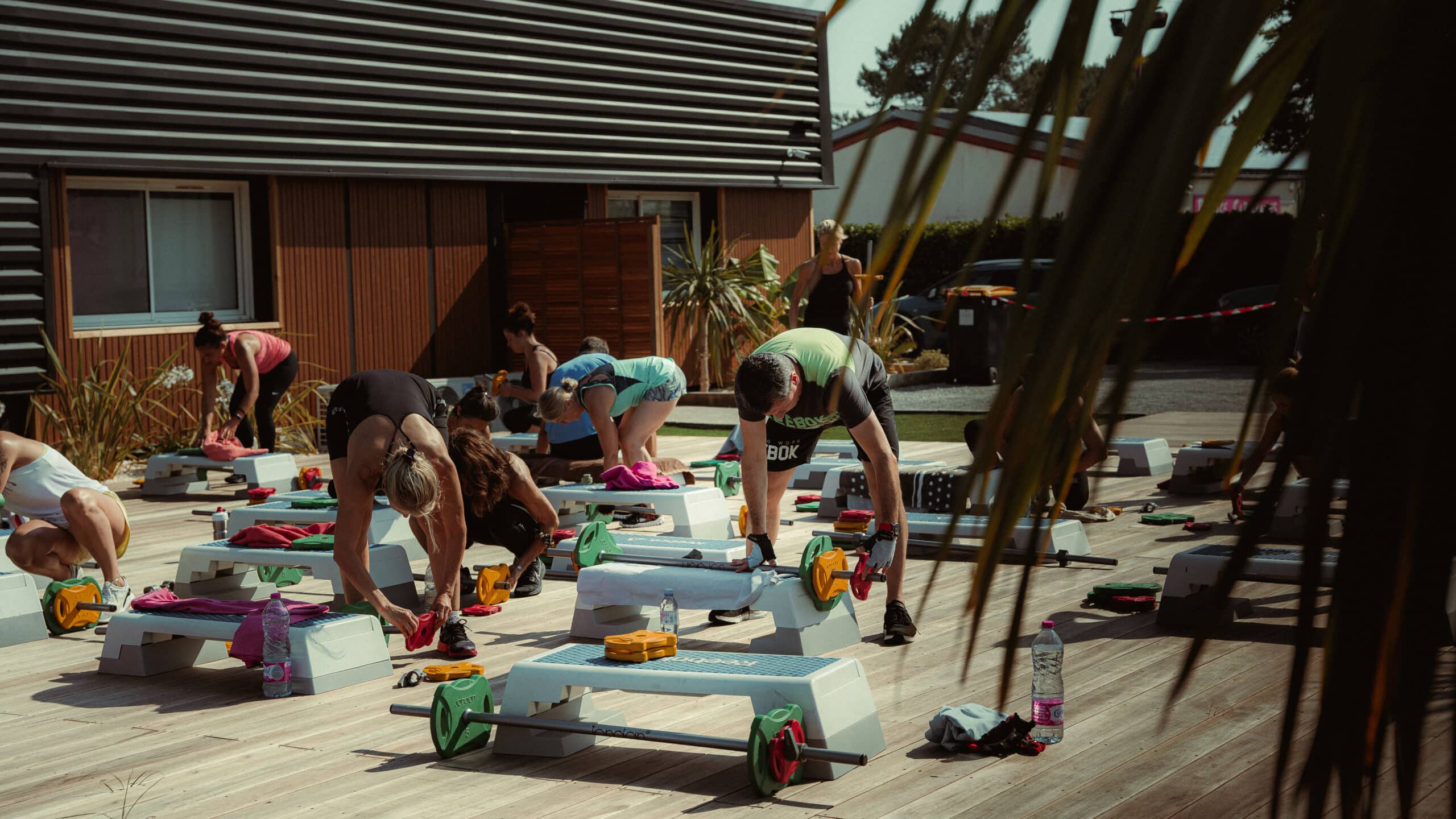 Séance de fitness collectif en extérieur avec haltères et steps sur une terrasse en bois ensoleillée.