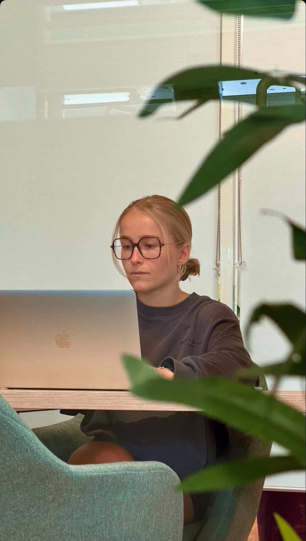Femme à lunettes concentrée travaillant sur son MacBook dans un bureau moderne entourée de plantes.