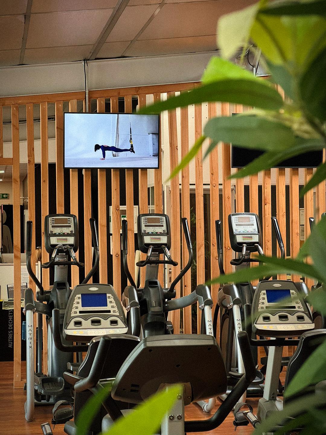 Vélos elliptiques Matrix dans une salle de sport moderne avec mur en bois, écran fitness et plantes.