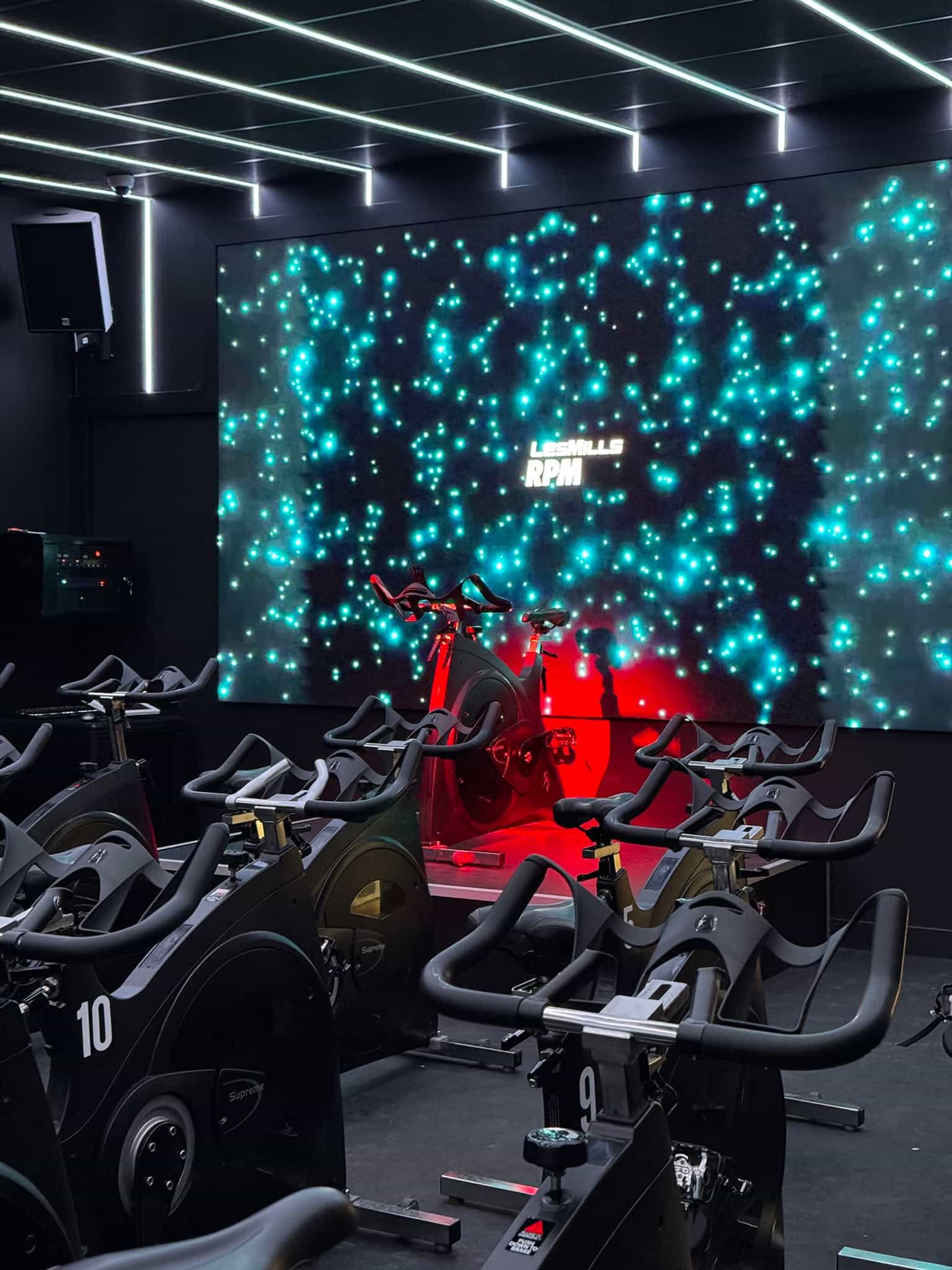 Studio de cycling Les Mills RPM avec vélos stationnaires devant un écran géant lumineux et éclairage néon.